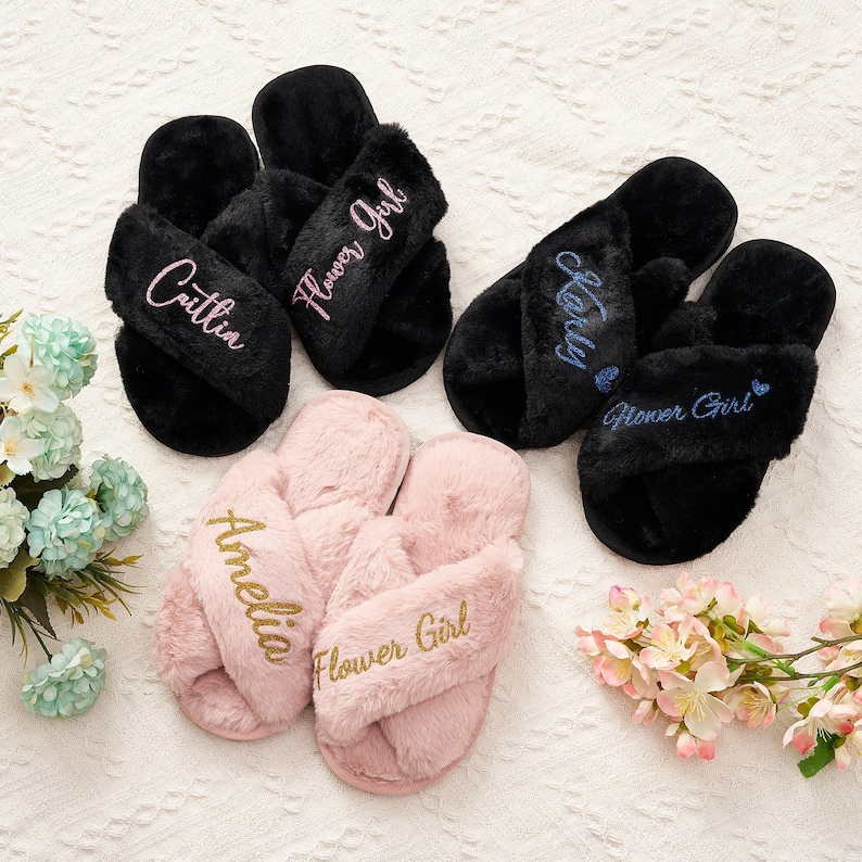 May include: Three pairs of fluffy slippers with the words "Flower Girl" embroidered on them. One pair is black with pink embroidery, one pair is black with blue embroidery, and one pair is pink with gold embroidery. The slippers are personalised with the names "Caitlin", "Carly", and "Amelia".