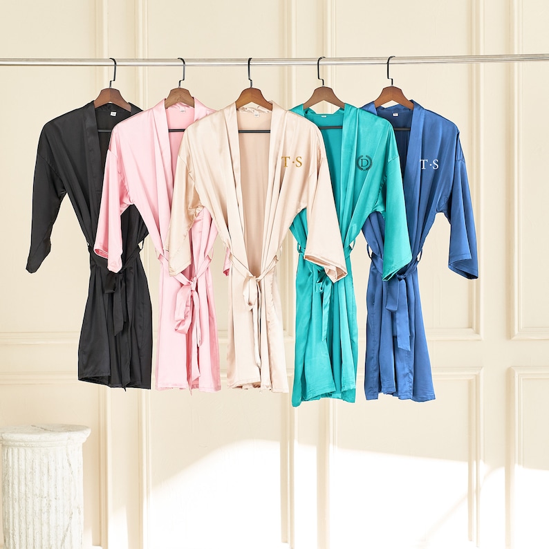 May include: Six satin robes in different colors hanging on a rack. The robes have embroidered initials on the left side of the chest. The robes are black, pink, beige, teal, and blue.