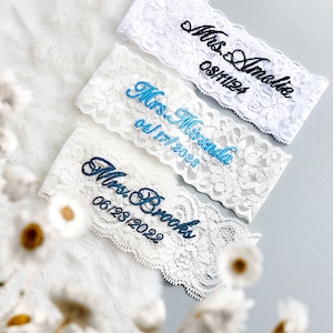 May include: Three white lace garters with embroidered text. The top garter reads "Mrs. Amelia" and "08/11/24". The middle garter reads "Mrs. Miranda" and "04/17/2023". The bottom garter reads "Mrs. Brooks" and "06/28/2022".