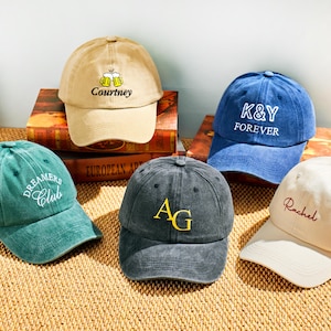 May include: Five baseball caps in various colors, including tan, blue, green, gray, and off-white. Each cap features embroidered text or a design, such as "Courtney," "K&Y FOREVER," "Dreamers Club," "AG," and "Rachel."