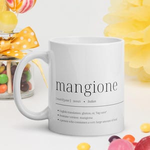 May include: White ceramic mug with a black design that reads "mangione | mangione | noun | Italian | English translation: glutton, or "big eater" | feminine version: mangiona | a person who consumes a very large amount of food"
