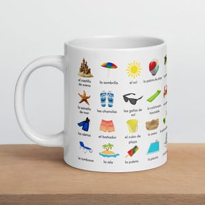 Pode incluir: Caneca de cerâmica branca com ilustrações com tema de praia e vocabulário espanhol. O design inclui um castelo de areia, estrela do mar, óculos de sol e outros itens essenciais de praia.