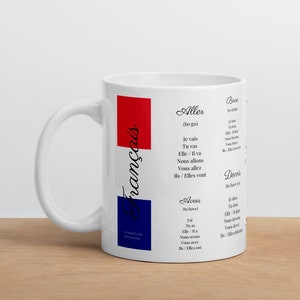 Könnte beinhalten: Eine weiße Keramiktasse mit einem rot-weiß-blauen Design. Die Tasse zeigt das Wort "français" in schwarzer Schrift und eine Liste französischer Verben in schwarzer Schrift auf weißem Hintergrund.
