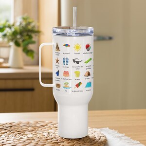 May include: A white travel mug with a handle and a clear lid, featuring a straw. The mug is adorned with colorful illustrations and French vocabulary related to the beach, including a sandcastle, sunglasses, and a beach ball.