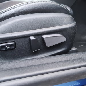 May include: Close-up of a black leather car seat with grey plastic controls. The controls include a button with a seat icon and two rectangular buttons.
