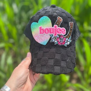 May include: Black mesh trucker hat with a pink heart, a horseshoe, and floral embroidery. The heart has a rainbow glitter finish and the word "boujee" is written on it.