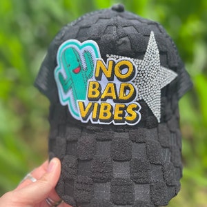 May include: Black trucker hat with a checkered pattern. The hat has a patch with a cartoon cactus and the text "NO BAD VIBES". A rhinestone star is on the side of the hat.