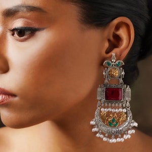 Könnte beinhalten: Silberne und goldfarbene Ohrringe mit roten und grünen Edelsteinen. Die Ohrringe haben ein Halbmond-Design mit Pfauen und Perlen-Akzenten.