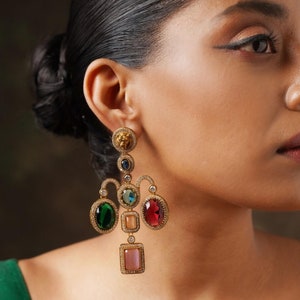 Op de afbeelding: Gouden chandelier oorbellen met een leeuwenkop charme, versierd met verschillende gekleurde edelstenen, waaronder groen, blauw, rood en roze.