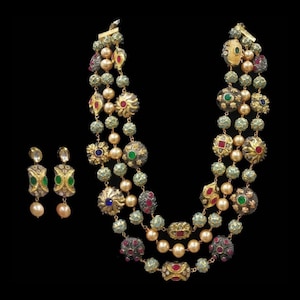 Op de afbeelding: Een set van een ketting met drie strengen en bijpassende oorbellen. De ketting heeft goudkleurig metaal met groene, rode en blauwe edelstenen en crèmekleurige parels. De oorbellen hebben een vergelijkbaar ontwerp.