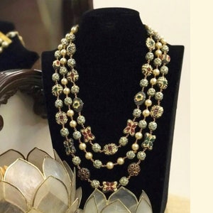 Peut inclure: Collier à plusieurs rangs présenté sur un mannequin noir. Le collier est composé de perles dorées, de fausses perles et d'éléments décoratifs avec des accents rouges, verts et dorés. Un bijou de déclaration.