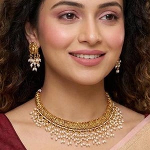 May include: A woman wearing a gold-toned necklace and matching earrings. The necklace is adorned with small pearls. She is wearing a maroon blouse and a light pink sari with a gold border.