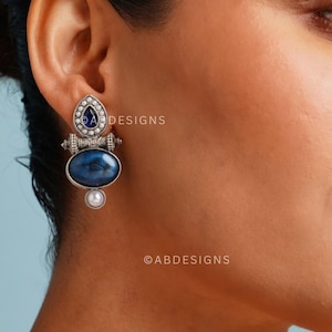 Può includere: Orecchini d'argento con una pietra blu e una perla pendente. Gli orecchini hanno una pietra blu a forma di lacrima nella parte superiore, una pietra blu ovale più grande al centro e una perla pendente nella parte inferiore.