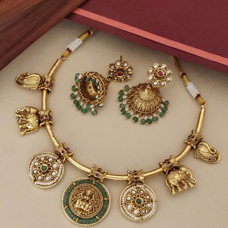 May include: Gold-toned jewelry set featuring a necklace with elephant and medallion charms, and matching earrings. The necklace has a curved design with intricate details. The earrings are bell-shaped with green beads. The medallions have green and white accents.