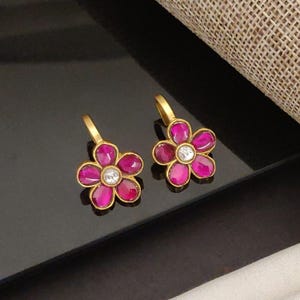 Puede incluir: Un par de pendientes de flores dorados. Cada pendiente presenta cinco pétalos de color magenta que rodean una piedra central clara y redonda. Los pendientes tienen un diseño de gancho simple para orejas perforadas.