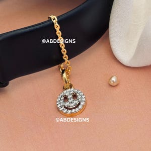 May include: A gold necklace featuring a smiley face pendant adorned with clear crystals. The pendant is suspended from a fine chain. A small pearl and the text "@ABDESIGNS" are also visible in the image.
