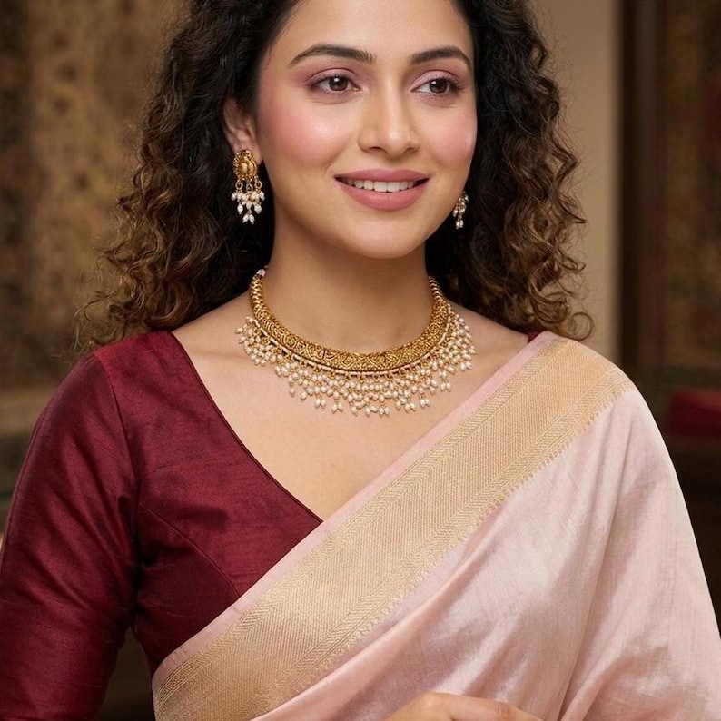 May include: A woman wearing a gold-toned necklace and matching earrings. The necklace is adorned with small pearls. She is wearing a maroon blouse and a light pink sari with a gold border.