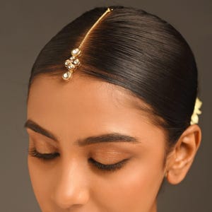 May include: A gold-coloured maang tikka, a traditional Indian forehead ornament, with a fine chain and a cluster of clear, teardrop-shaped gemstones. The chain extends to the hairline, and the gemstones sit on the forehead.