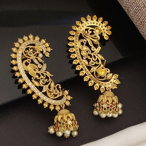 May include: A pair of gold-colored ear cuffs with intricate floral and paisley designs. The earrings feature small, dangling bells adorned with pearls and colorful gemstones. The ear cuffs are embellished with clear crystals and green and red gemstones.