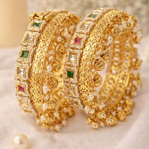 May include: Two gold bangles featuring detailed floral designs, small white pearls, and colourful gemstones. The bangles have a textured finish and are decorated with dangling gold charms. The overall design is ornate and elegant.