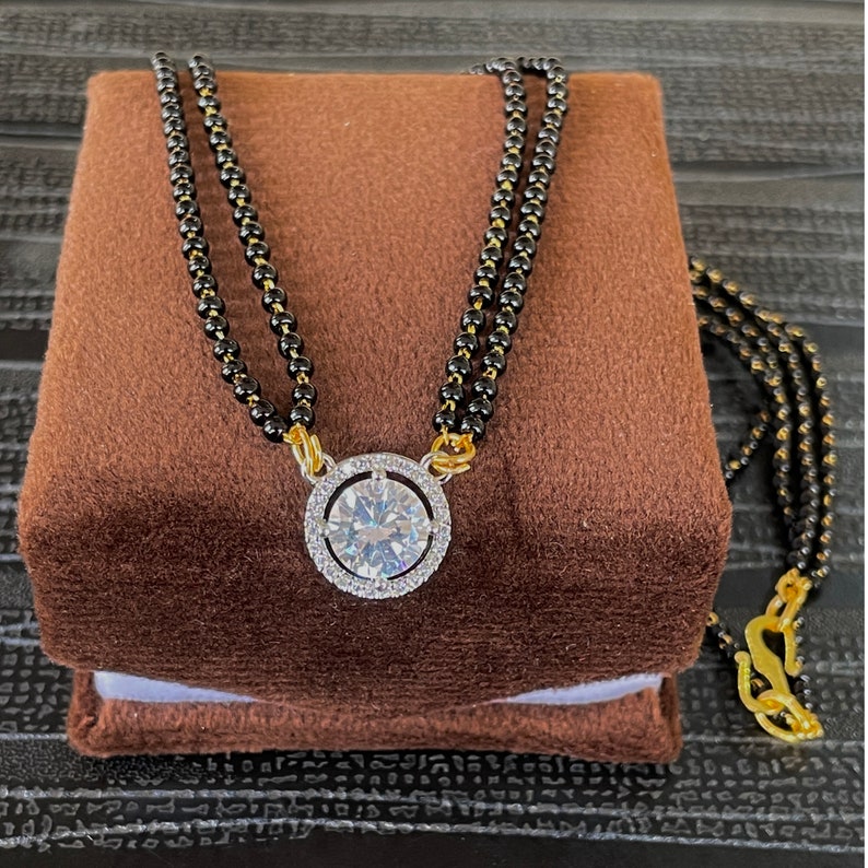 May include: A black beaded necklace with a gold pendant featuring a large, clear, round gemstone surrounded by smaller, clear gemstones.