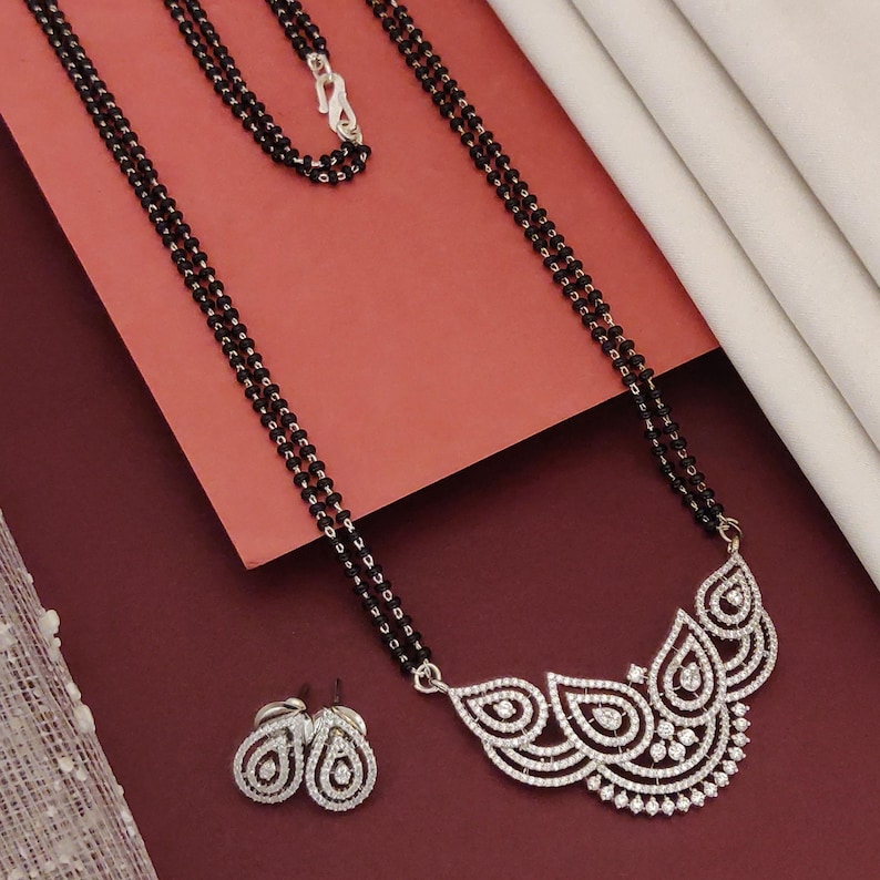 Puede incluir: Un collar de cuentas negras con un cierre plateado y un colgante con un dise&ntilde;o blanco y ornamentado. Tambi&eacute;n hay pendientes a juego con un dise&ntilde;o similar. Las joyas se exhiben sobre una superficie roja.