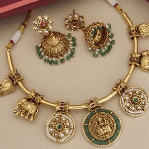 May include: A gold-toned jewelry set featuring a necklace and earrings. The necklace has elephant and medallion pendants with green and white accents. The earrings are ornate, with green bead dangles. The set has red and clear stone embellishments.