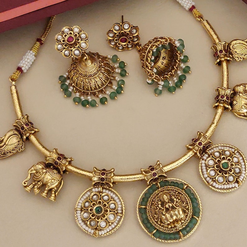 May include: A gold-toned jewelry set featuring a necklace and earrings. The necklace has elephant and medallion pendants with green and white accents. The earrings are ornate, with green bead dangles. The set has red and clear stone embellishments.