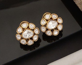 May include: A pair of gold-toned stud earrings. Each earring features a central, large, clear gemstone surrounded by smaller, clear gemstones. The earrings are set in a gold-colored metal with a floral design.