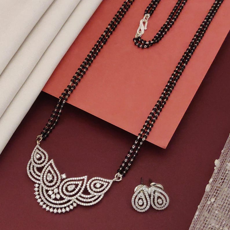 Puede incluir: Un collar de cuentas negras con un cierre plateado y un colgante con un dise&ntilde;o blanco adornado con piedras claras incrustadas. Tambi&eacute;n hay pendientes a juego con un dise&ntilde;o similar. El conjunto de joyas se exhibe sobre una superficie roja.