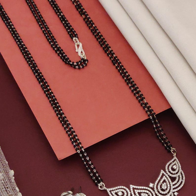 Puede incluir: Un collar de cuentas negras con un cierre plateado y un colgante decorativo. El colgante presenta un dise&ntilde;o floral con piedras claras y brillantes. El collar se exhibe sobre una superficie roja con una tela blanca en el fondo.