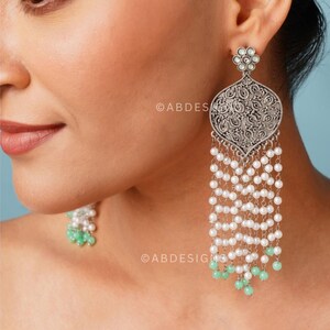 Peut inclure: Boucles d'oreilles pendantes argentées avec un motif filigrané orné. Les boucles d'oreilles présentent un pendentif en forme de goutte d'eau avec des motifs complexes, orné de rangées de perles blanches et de perles vert menthe. Le haut a un motif floral.