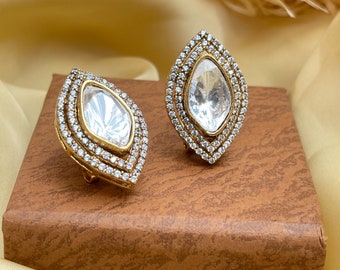 May include: A pair of gold-toned stud earrings. Each earring features a large, clear, teardrop-shaped center stone, surrounded by a halo of smaller, clear stones. The earrings are displayed on a brown surface against a soft, yellow background.