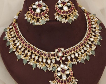 May include: A gold-toned necklace and earring set. The necklace features a series of red and white stones, with green and white bead accents. The earrings have a similar design, with a circular shape and matching stones and beads. The jewelry is displayed on a brown surface.