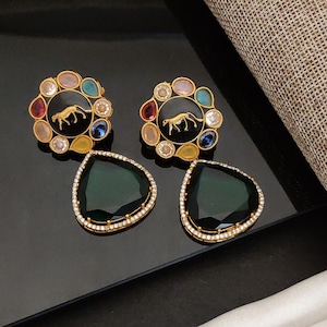 May include: A pair of gold-toned earrings with a black center featuring a cheetah design, surrounded by colorful gemstones. Below, two large, dark green, teardrop-shaped earrings with a diamond-like border.