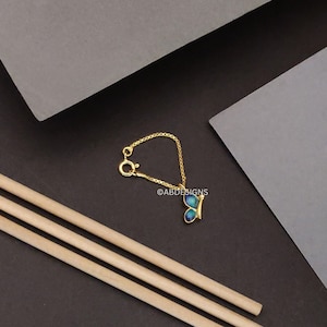 May include: A gold-colored butterfly charm with blue and green enamel wings, attached to a delicate chain. The charm is connected to a clasp. The image is set against a dark background with wooden dowels and gray paper.