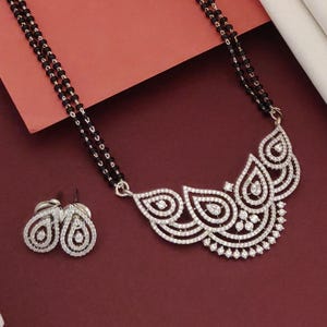 Puede incluir: Un conjunto de collar y pendientes plateados. El collar presenta una cadena de cuentas negras y un colgante con un dise&ntilde;o curvo detallado incrustado con piedras preciosas transparentes. Los pendientes tienen forma de l&aacute;grima, tambi&eacute;n con piedras preciosas transparentes.