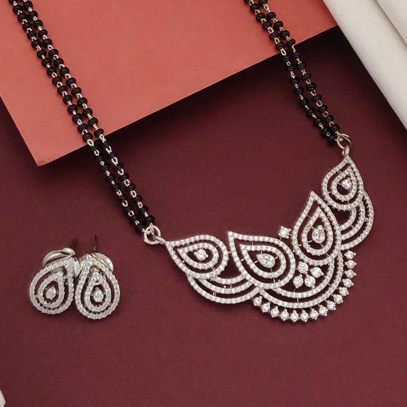 Puede incluir: Un conjunto de collar y pendientes plateados. El collar presenta una cadena de cuentas negras y un colgante con un dise&ntilde;o curvo detallado incrustado con piedras preciosas transparentes. Los pendientes tienen forma de l&aacute;grima, tambi&eacute;n con piedras preciosas transparentes.