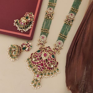 Può includere: Un set di gioielli color oro con una lunga collana e orecchini abbinati. La collana ha più fili di perline verdi, dettagli dorati ornati e un grande ciondolo con pietre preziose rosse e verdi. Gli orecchini hanno un design simile.