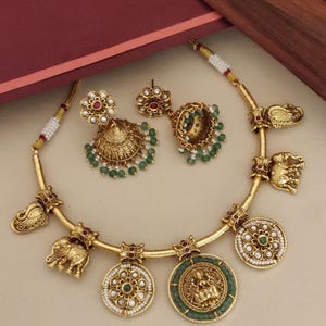 May include: A gold-toned jewelry set featuring a necklace and matching earrings. The necklace has elephant and peacock charms, and circular pendants with floral and deity designs. The earrings are bell-shaped with green and white beads, and the necklace has similar bead accents.