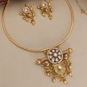 May include: A gold choker necklace with a large, ornate pendant featuring a circular design with white stones. The pendant has intricate details and dangling accents. Matching earrings with a similar design are included.