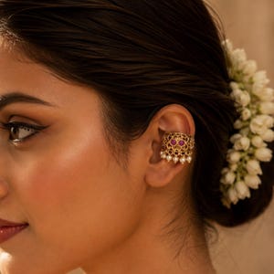 Puede incluir: Un pendiente tipo ear cuff de tono dorado con un diseño de filigrana metálica intrincada, acentuado con dos piedras redondas de color magenta y una fila de pequeñas cuentas blancas similares a perlas colgando del borde inferior.