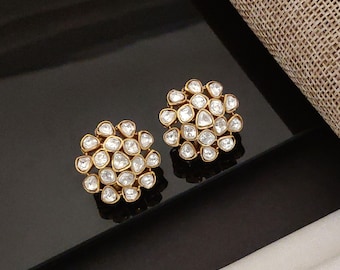 May include: A pair of gold-toned stud earrings. Each earring features a cluster of clear, irregularly shaped stones set in a circular arrangement. The earrings are displayed on a glossy black surface, with a neutral-toned fabric in the background.