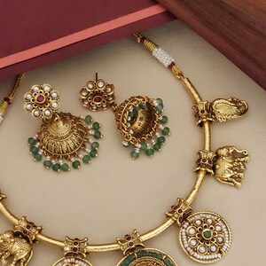 May include: A gold-toned jewelry set featuring a necklace and earrings. The necklace has elephant and floral motifs, with a central pendant. The earrings are bell-shaped with green beads and floral accents. The set has a vintage aesthetic.