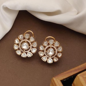 May include: A pair of gold-coloured stud earrings. Each earring has a central, large, clear, faceted stone surrounded by smaller, clear, triangular-shaped stones. The earrings are set against a brown background with white fabric.