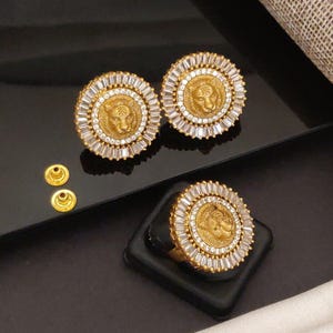 Può includere: Set di gioielli color oro con un anello e due paia di orecchini. Ogni pezzo presenta una testa di leone al centro, circondata da cristalli trasparenti, rettangolari e rotondi. Gli orecchini e l'anello sono esposti su una superficie nera.