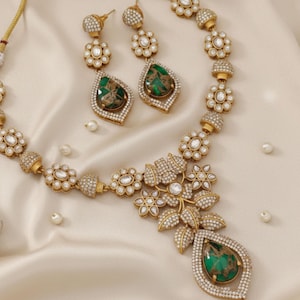 Peut inclure: Un ensemble de bijoux doré comprenant un collier et des boucles d'oreilles assorties. Le collier présente un motif floral avec des pierres claires et un grand pendentif en forme de goutte d'eau verte. Les boucles d'oreilles ont également des pierres vertes et un motif floral.