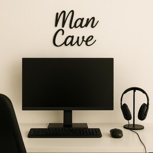 May include: A black computer monitor, keyboard, and mouse sit on a white desk. A pair of black headphones rests on a stand to the right. The words "Man Cave" are displayed on the wall above the monitor.
