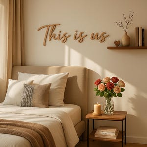 以下が含まれることがあります： ベッド、枕、そして「This is us」と書かれた木製の看板がある寝室のインテリア。バラの花瓶、ろうそく、本がサイドテーブルに置かれています。小さな棚には花瓶、本、ハートが置かれています。
