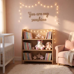May include: A light wood bookshelf filled with colourful books, star-shaped pillows, and stuffed animals. The wall above the shelf has the phrase "You are my sunshine" in wooden letters, and is decorated with string lights. A cot and armchair are also in the room.